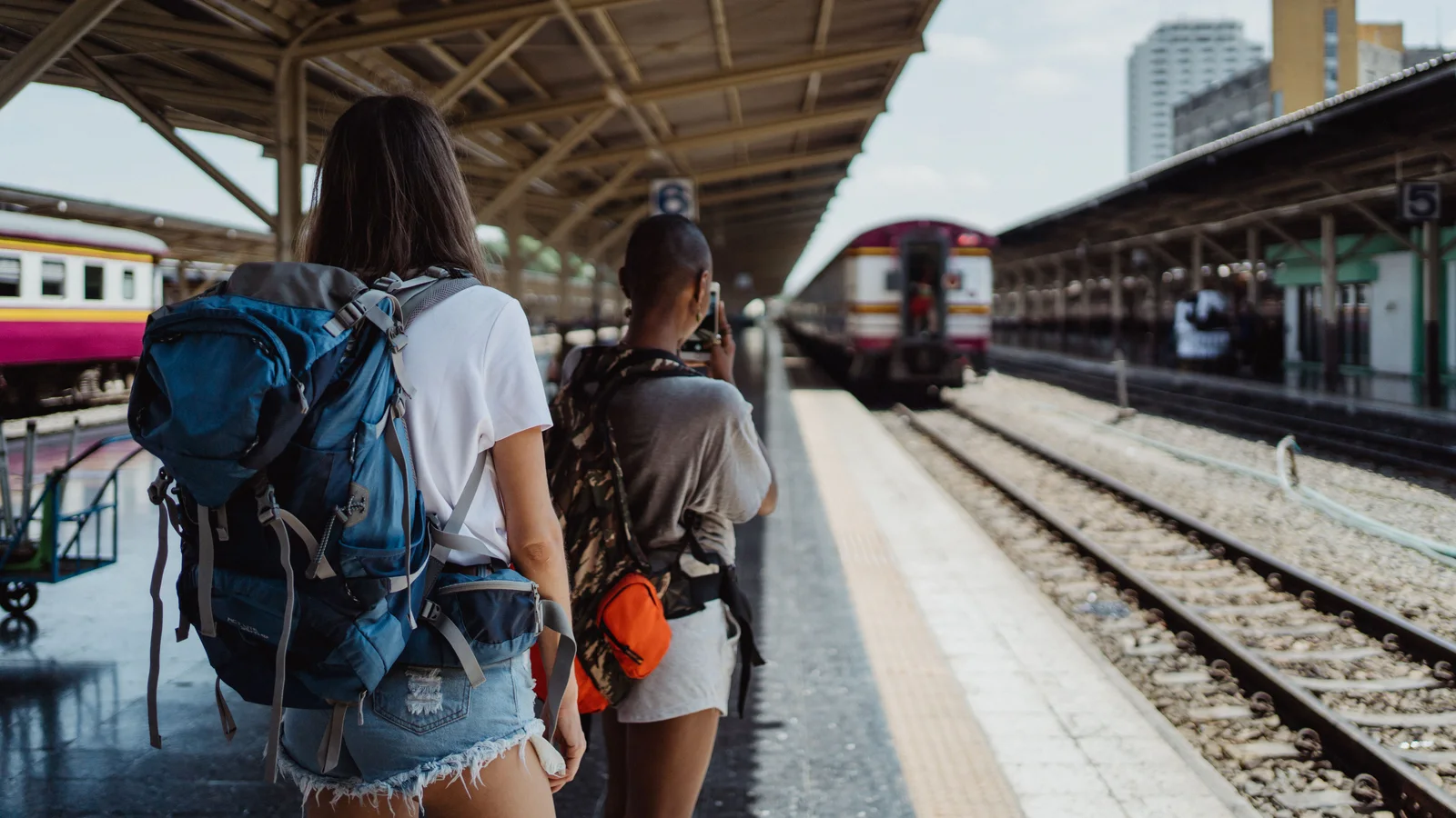 Viaggiatrici con zaini su una banchina ferroviaria in attesa del treno.